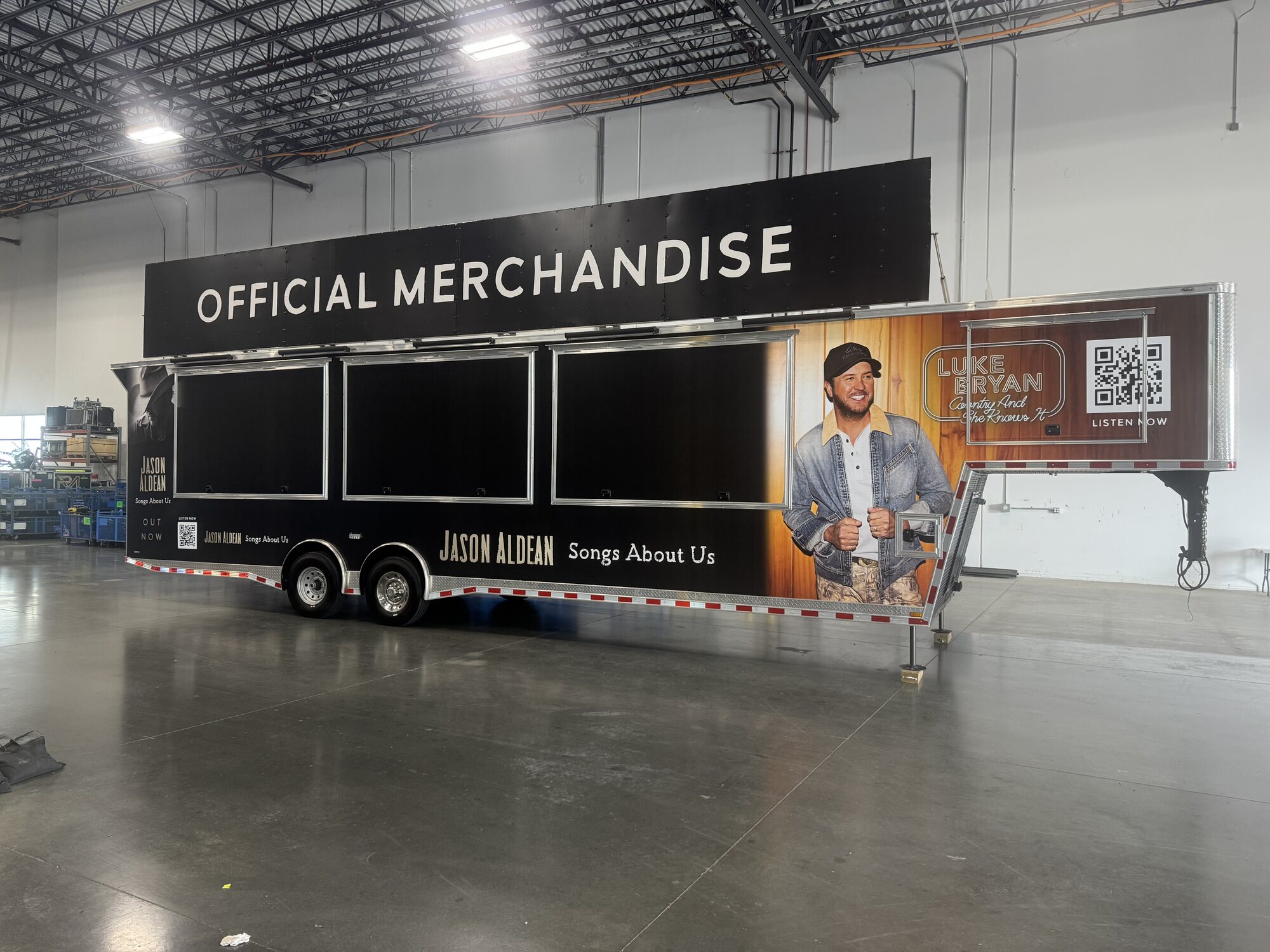 Luke Bryan and Jason Aldean official merchandise trailer with full black graphics, artist photo and QR codes, parked in a warehouse